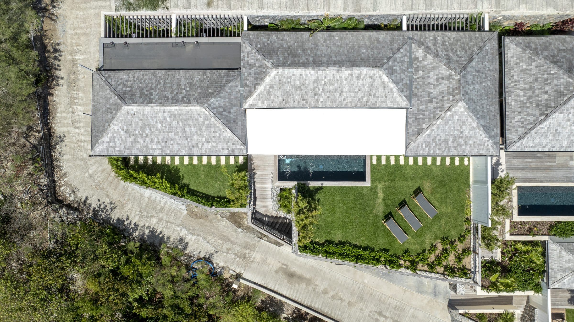 Straight-down drone view of the villa’s shingled roof pavilions, pool and lawn with three loungers