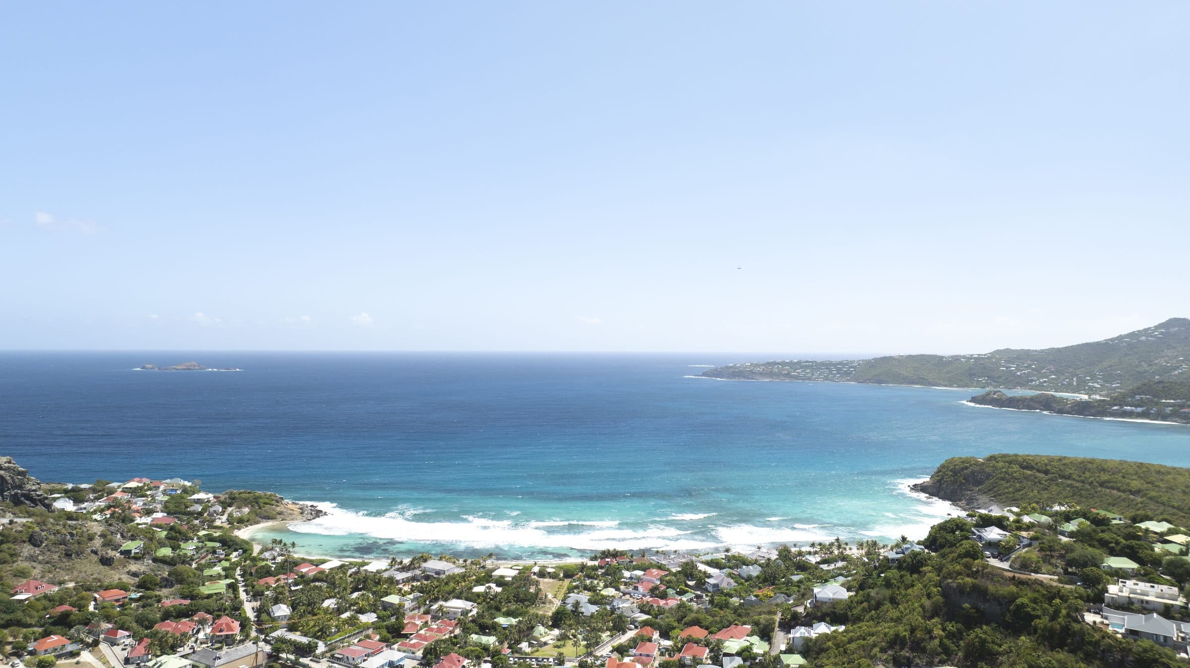 High-altitude aerial view of the full curve of Saint-Jean Bay with a small offshore islet and the village below
