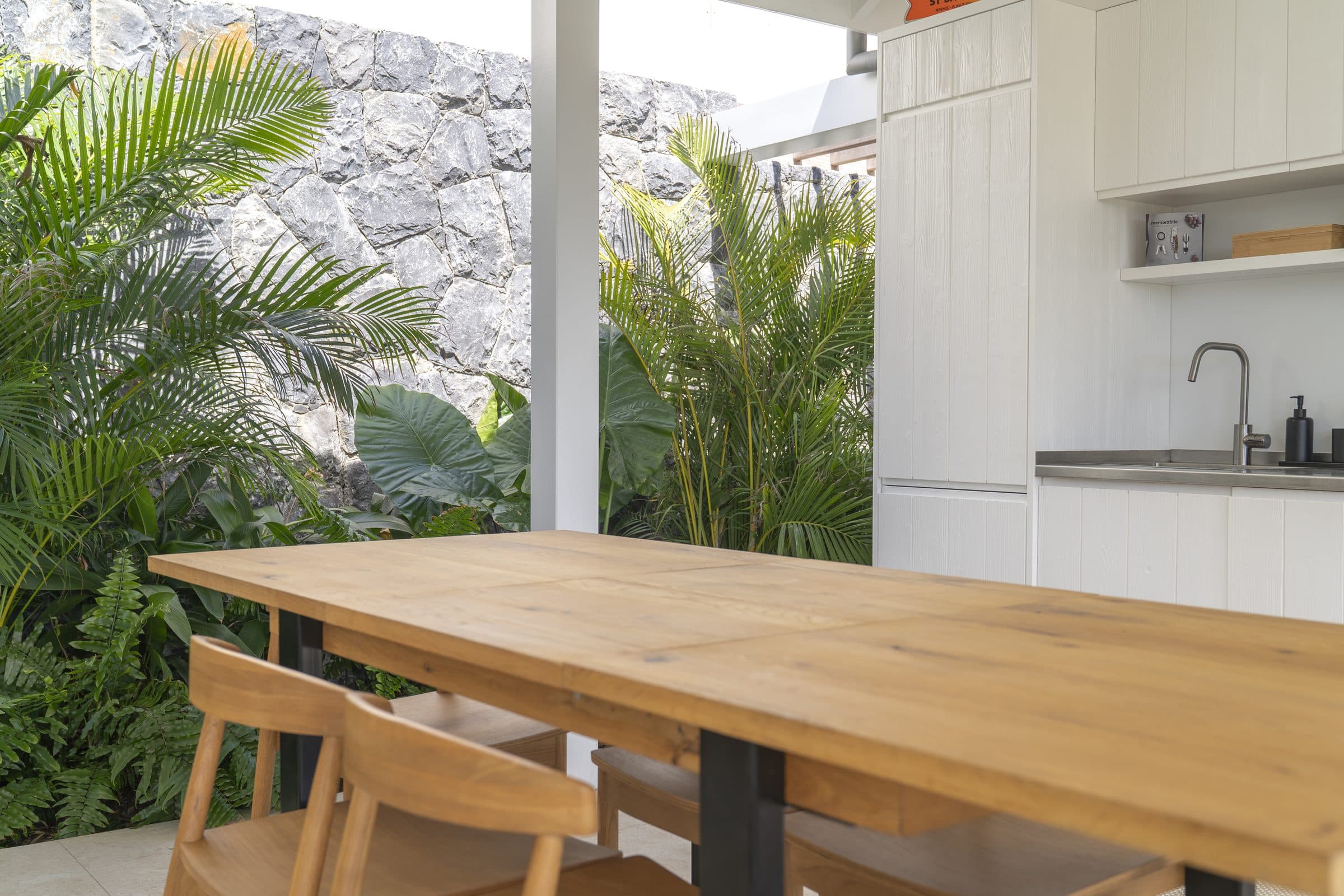 Solid oak dining table under the covered terrace, open kitchen and tropical planting against a stacked-stone wall