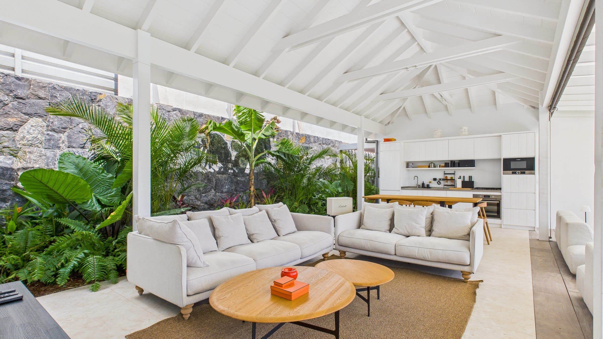 Open-air living pavilion with vaulted white ceiling, two linen sofas, round oak coffee tables and the open kitchen behind