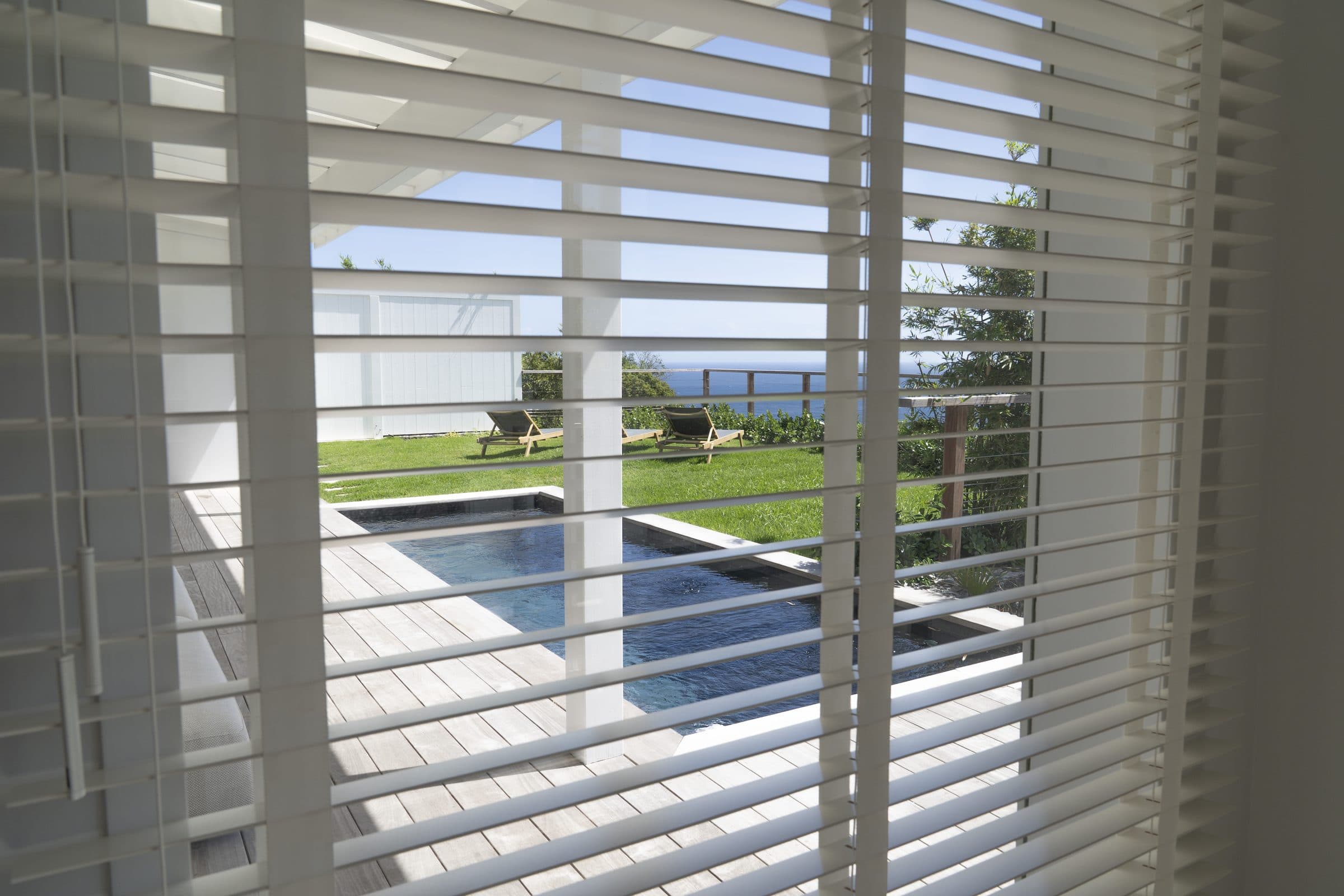 View through white louvre shutters onto the wooden pool deck, lawn loungers and the sea