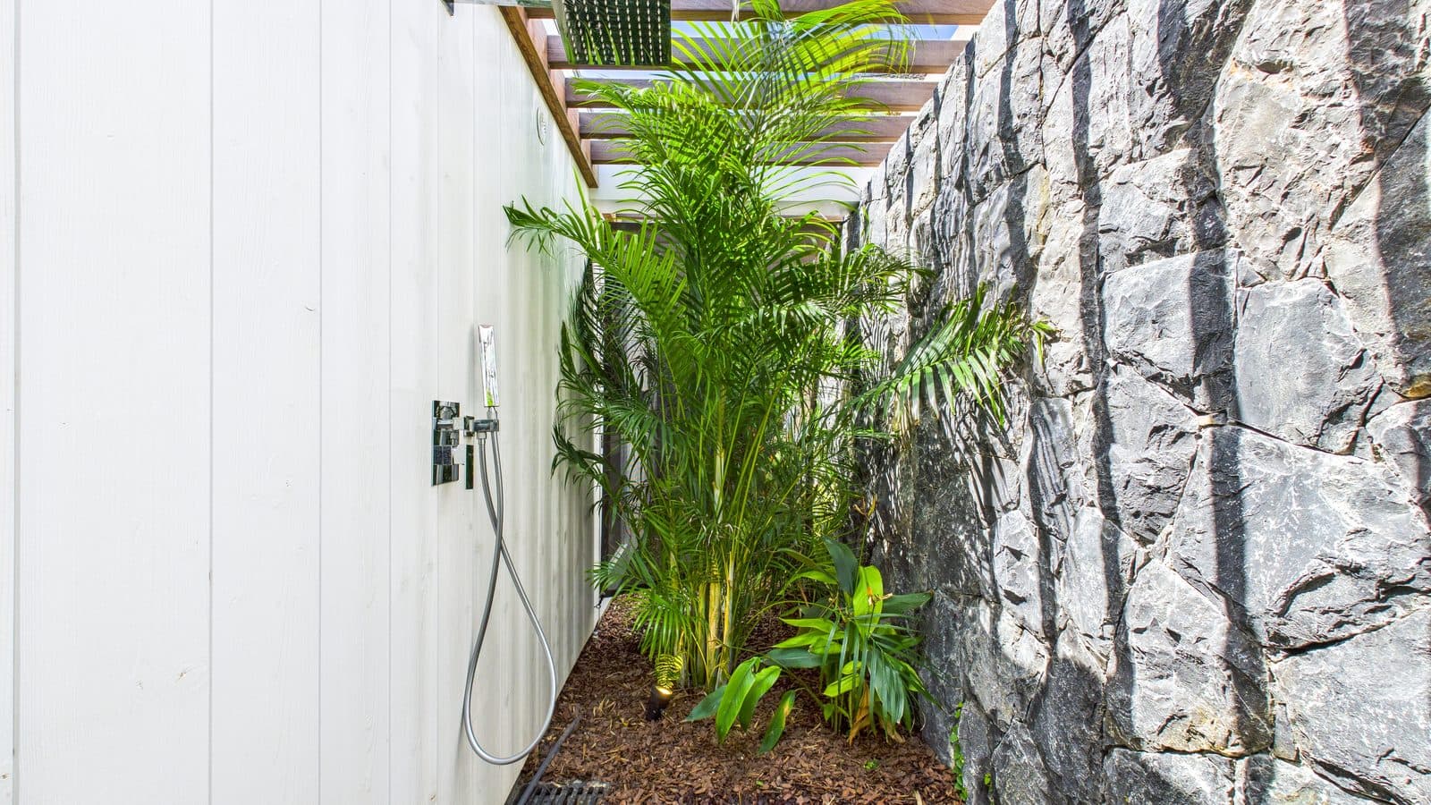 Open-air shower corridor between the villa’s timber wall and a stacked-stone wall, shaded by palms overhead