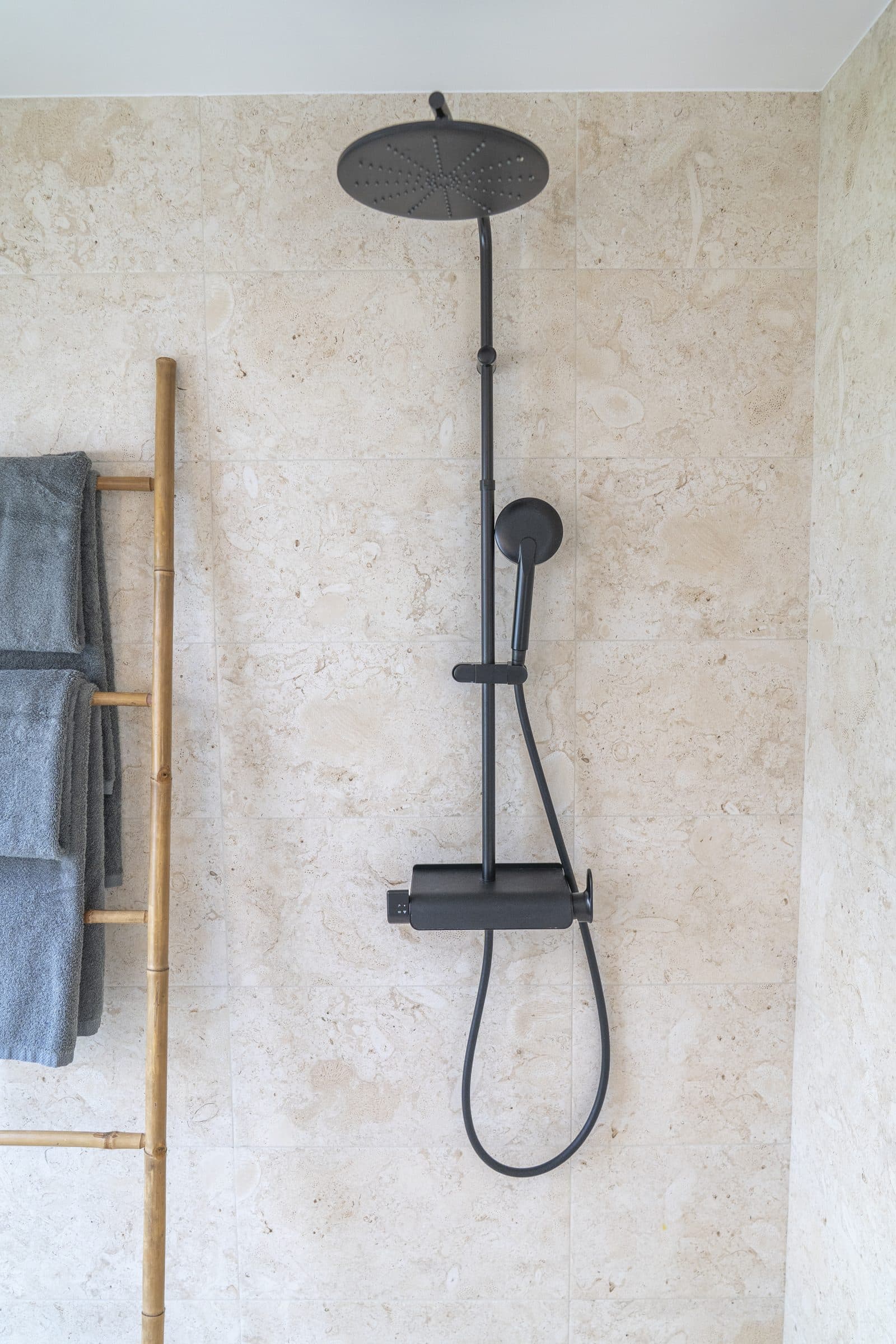 Matte black rain shower and handheld spray mounted on a travertine-tiled wall, bamboo towel ladder alongside