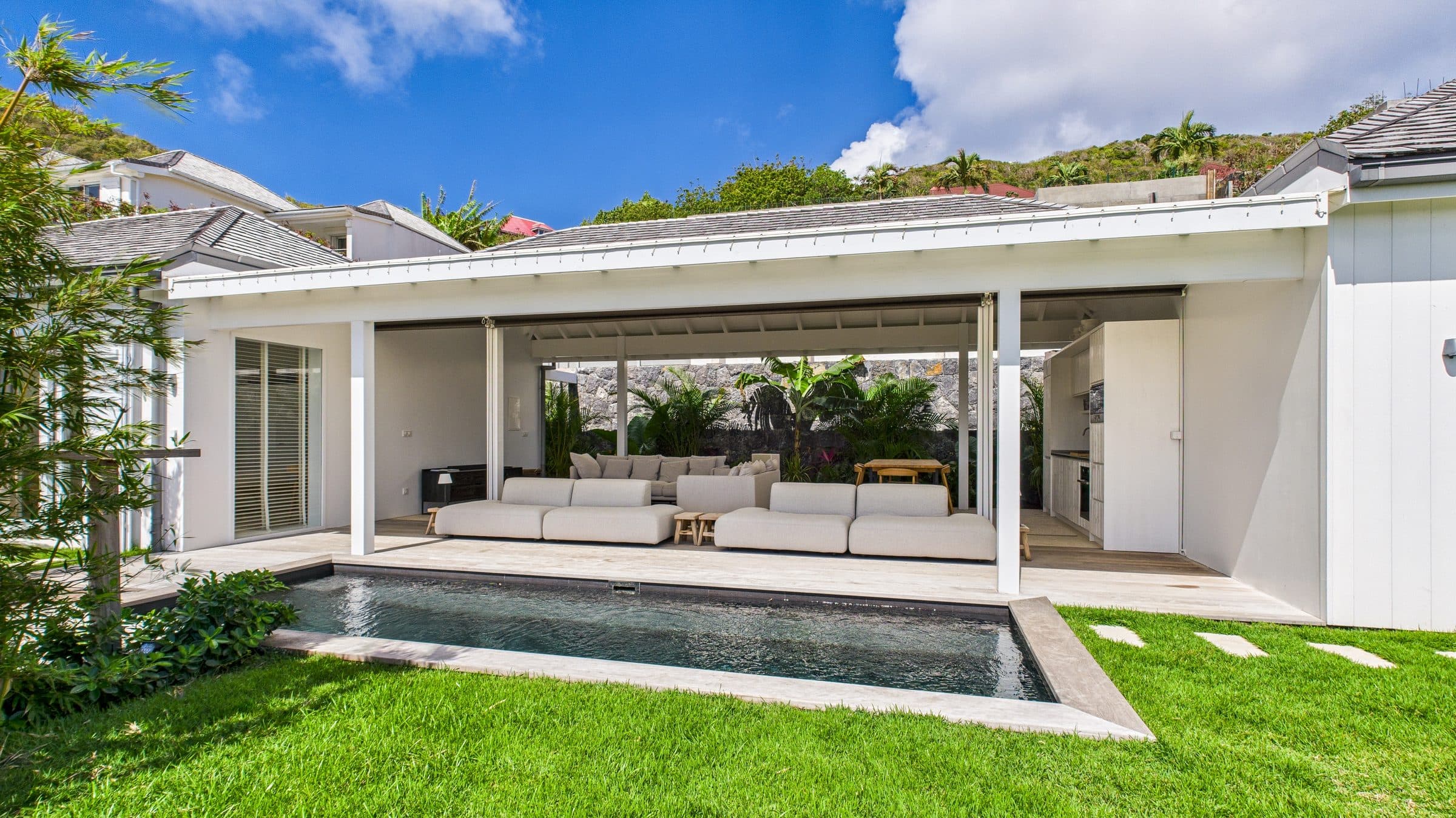 Front elevation of the villa’s living pavilion opening onto the pool and lawn, hills rising behind the roofline