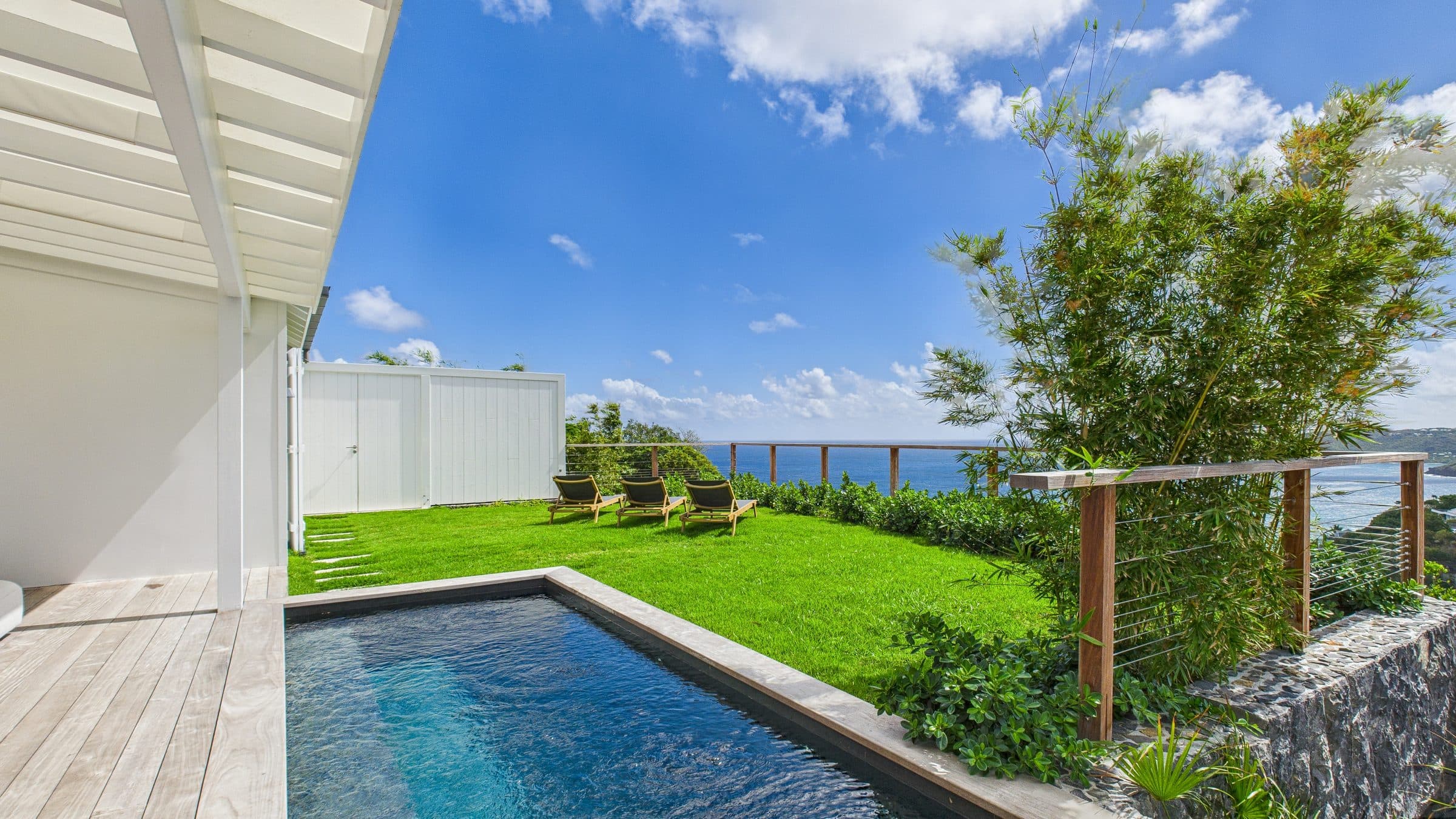 Poolside lawn with loungers under open sky, timber deck and the sea horizon beyond the cable railing
