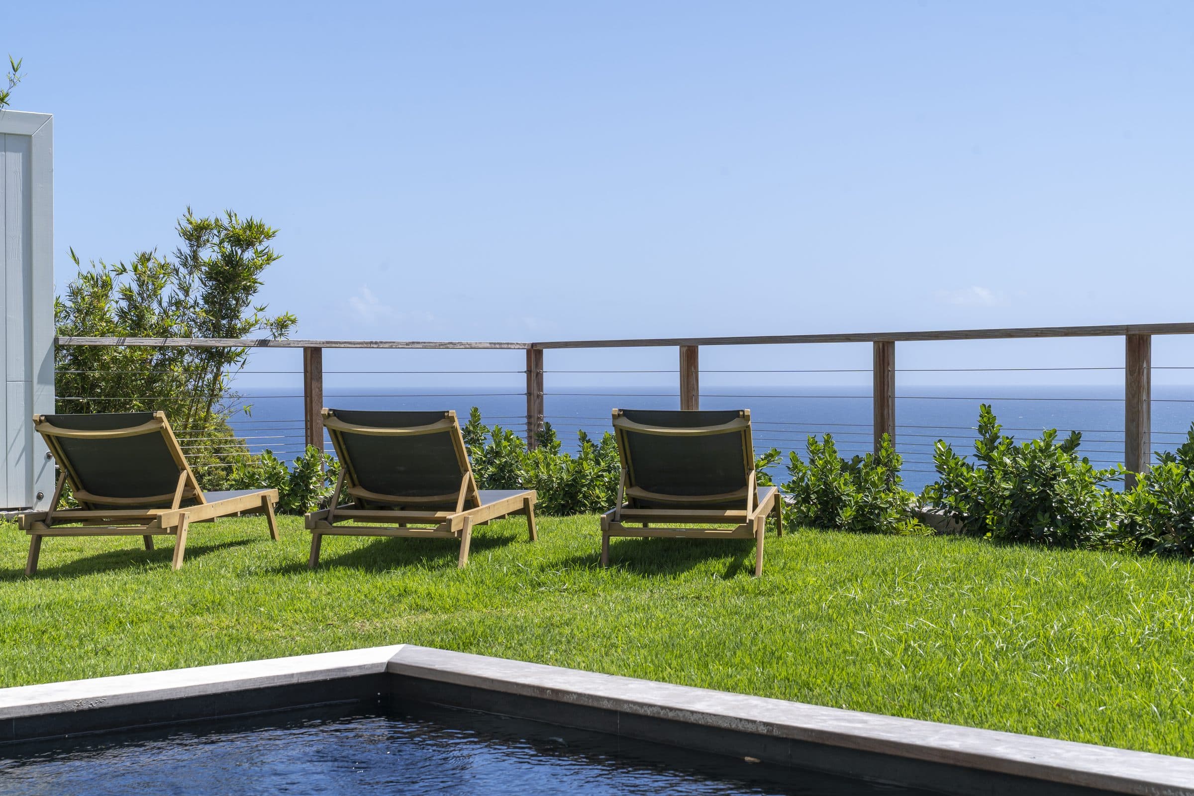 Dark-stone plunge pool bordered by lawn, three loungers facing the open sea