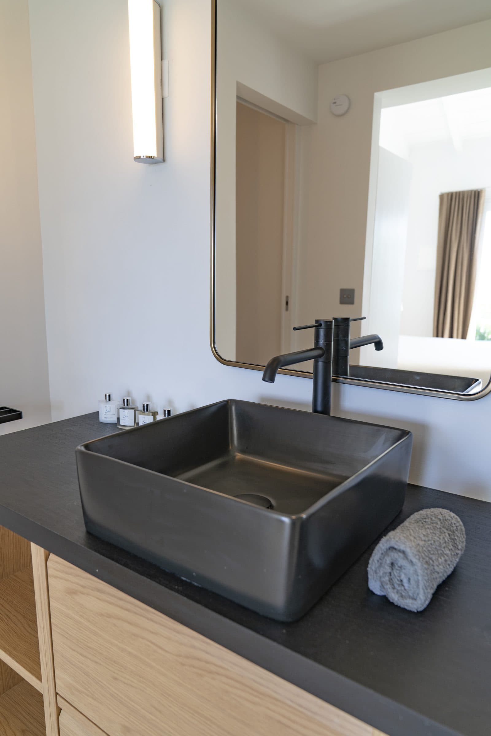 Square dark concrete vessel sink and matte black tap on an oak vanity, rolled towel and amenities set out